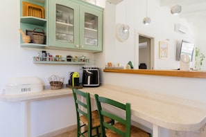 Een keuken met een broodmachine, een koffiezetapparaat en een broodrooster. Er staan groene stoelen bij een houten tafel.