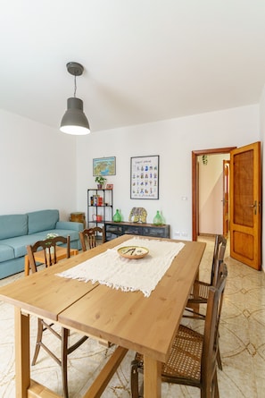 A dining area with a wooden table, chairs, a hanging light fixture, a blue sofa, a bookshelf, and a door.