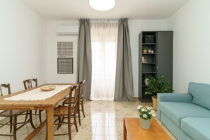 A dining area with a wooden table and chairs, a sofa, a bookshelf, and a potted plant.