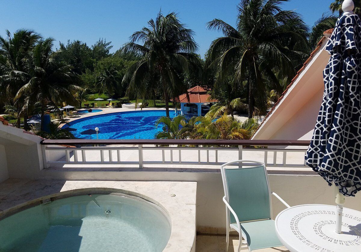 Zona de piscina con hidromasaje, sillones de descanso y vistas a una piscina más grande y palmeras.