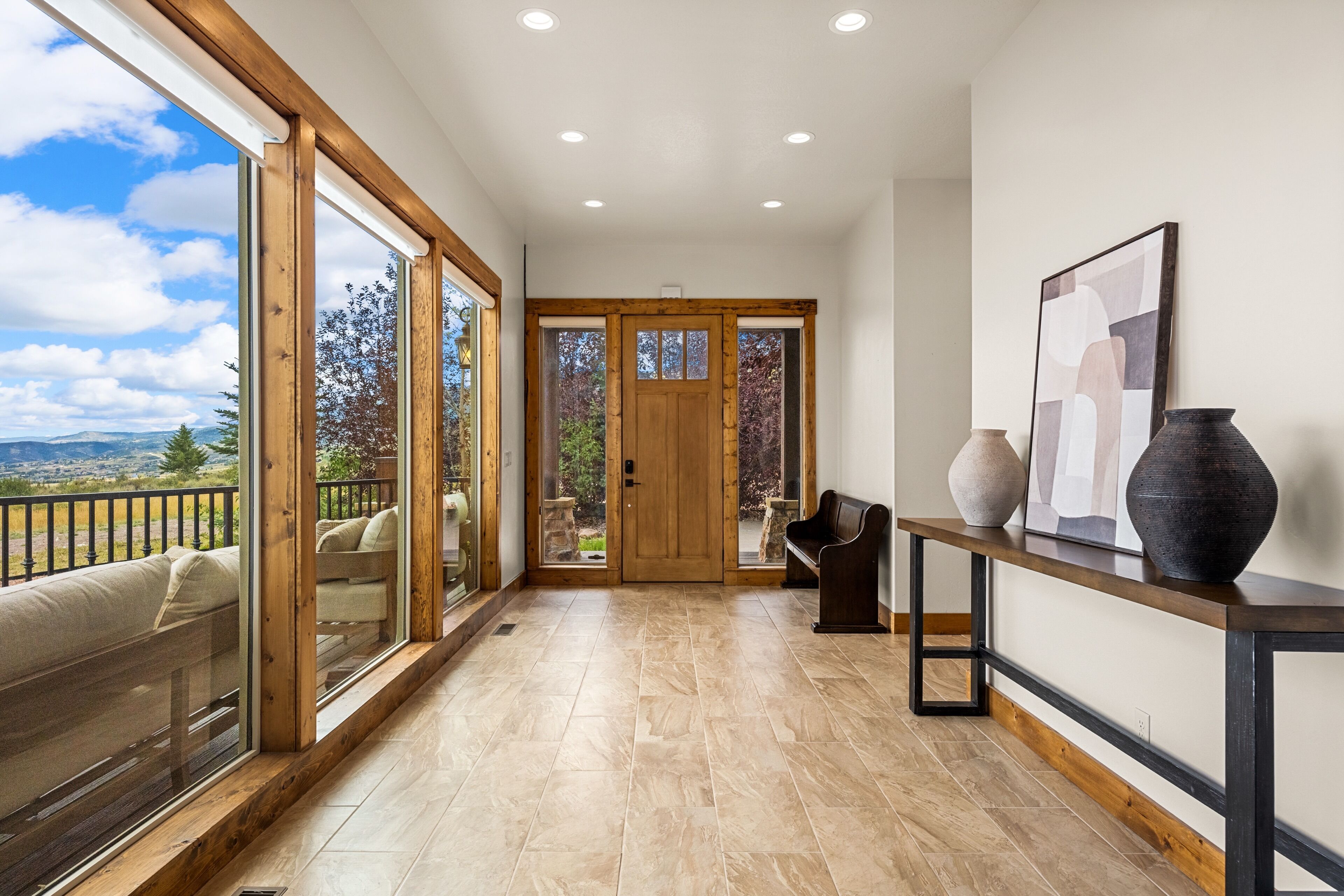 A spacious hallway with a view of the outdoors, a wooden bench, a vase, and a framed picture on a console table.