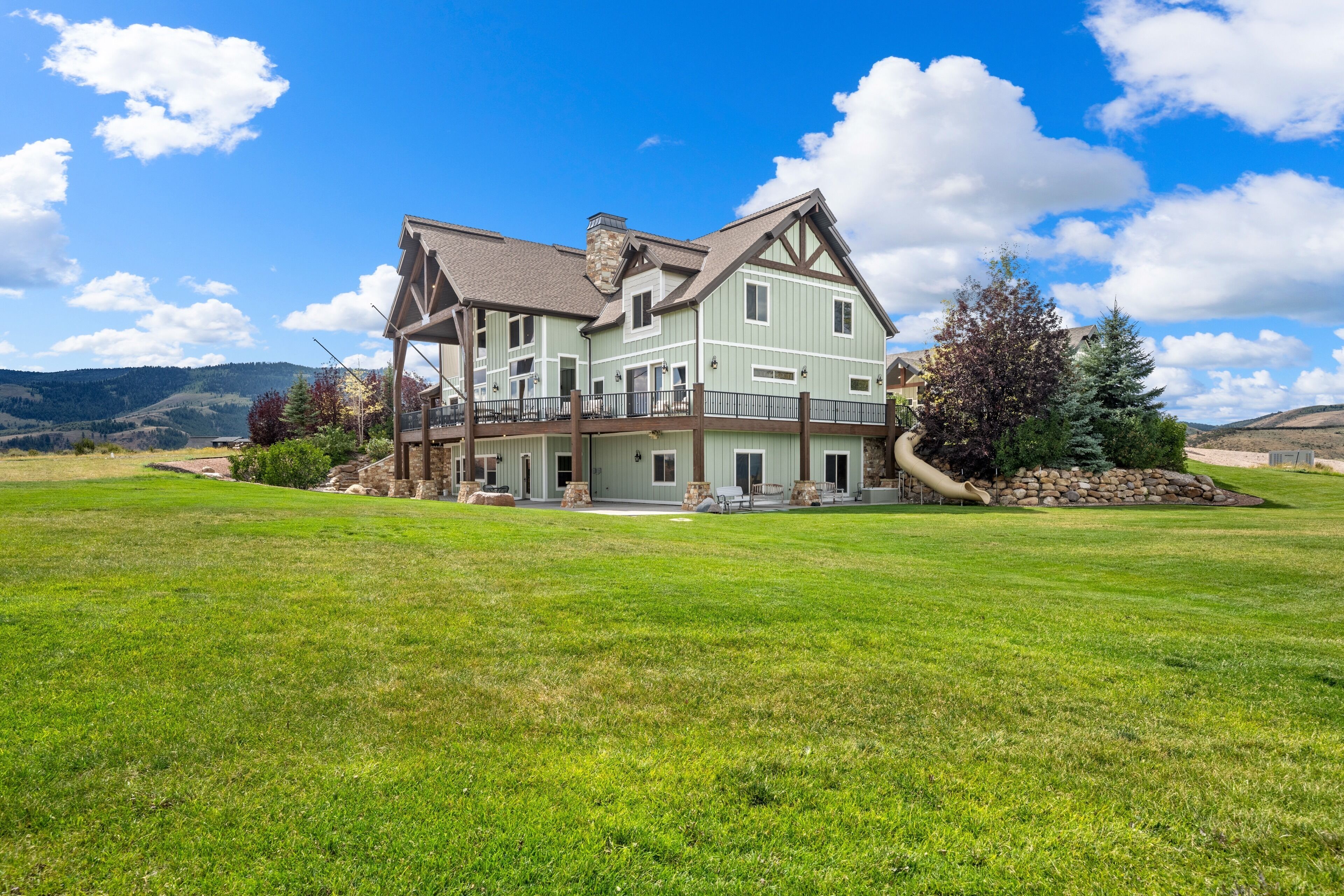 A large two-story house with a balcony, surrounded by a well-maintained lawn and a mountain backdrop.