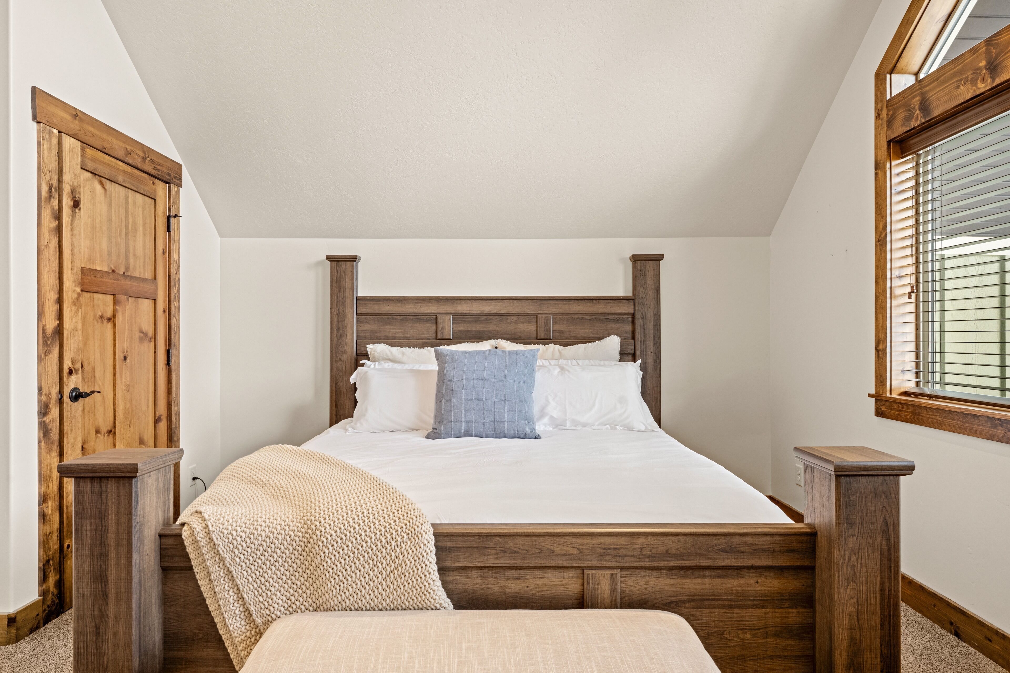 A bedroom with a wooden headboard, a bed with white linens, a knitted chair, and a window with blinds.