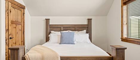 A bedroom with a wooden headboard, a bed with white linens, a knitted chair, and a window with blinds.