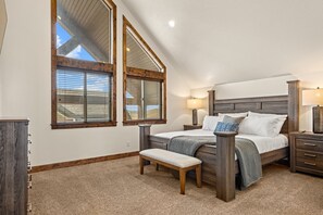 Un dormitorio con techo inclinado, una cama grande con cabecera de madera, dos lámparas de noche, un banco y una mesita de noche con lámpara.
