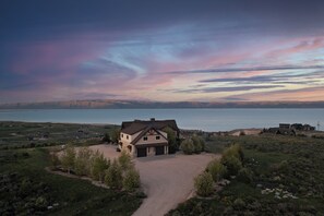 A large house with a driveway, surrounded by a natural landscape and a body of water in the background.