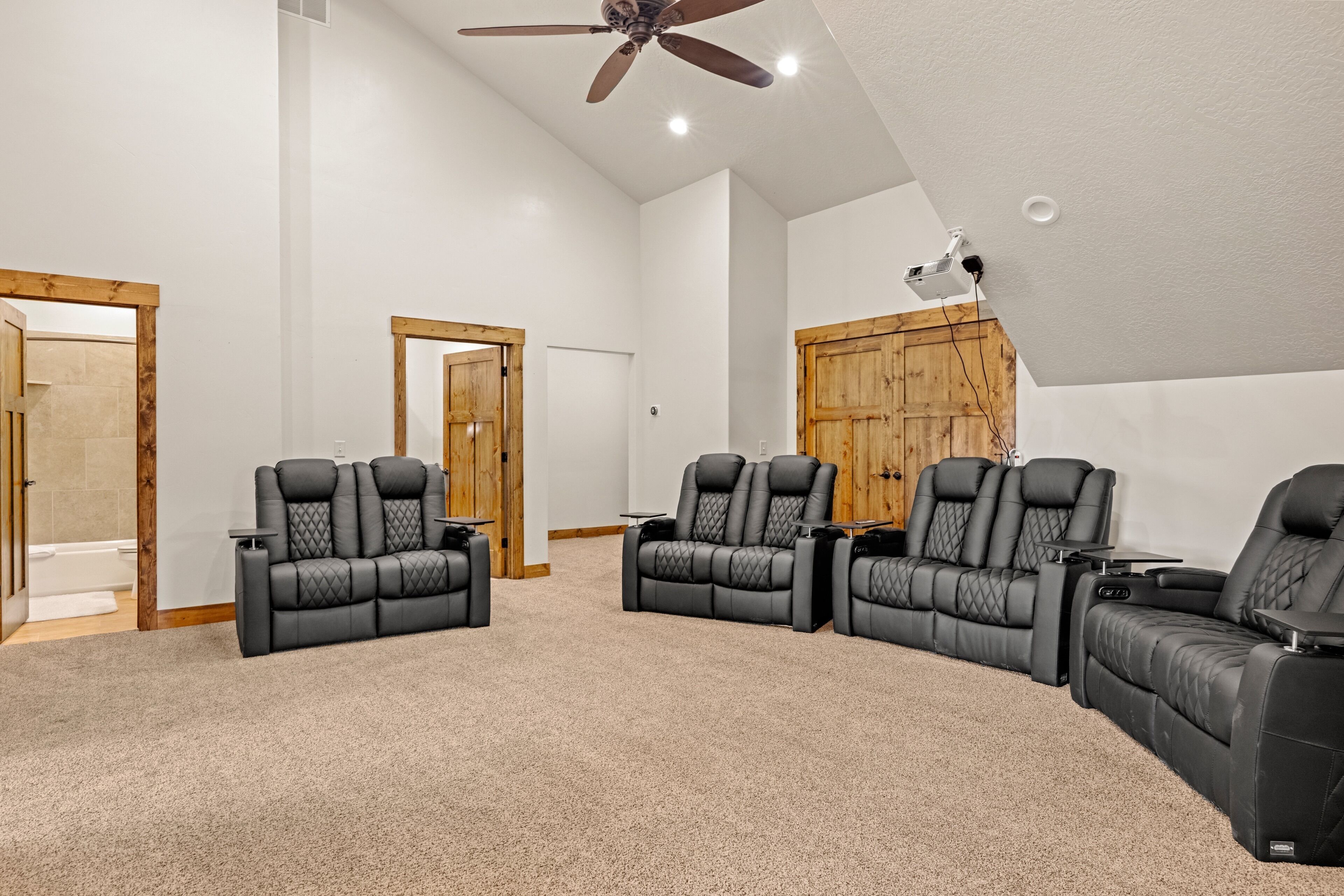 Una sala de cine en casa con tres sillones reclinables negros, un ventilador de techo y un proyector montado en la pared.