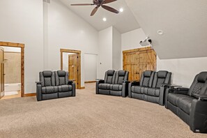 Sala de cinema com três poltronas reclináveis pretas, um ventilador de teto e um projetor fixado na parede.