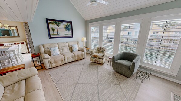 A living room with a beige sofa, a patterned armchair, and a grey armchair. There is a painting of a beach scene on the wall, and the room has large windows with blinds. - Sandpiper Cove 0013 3 Bedroom Villa (Destin)