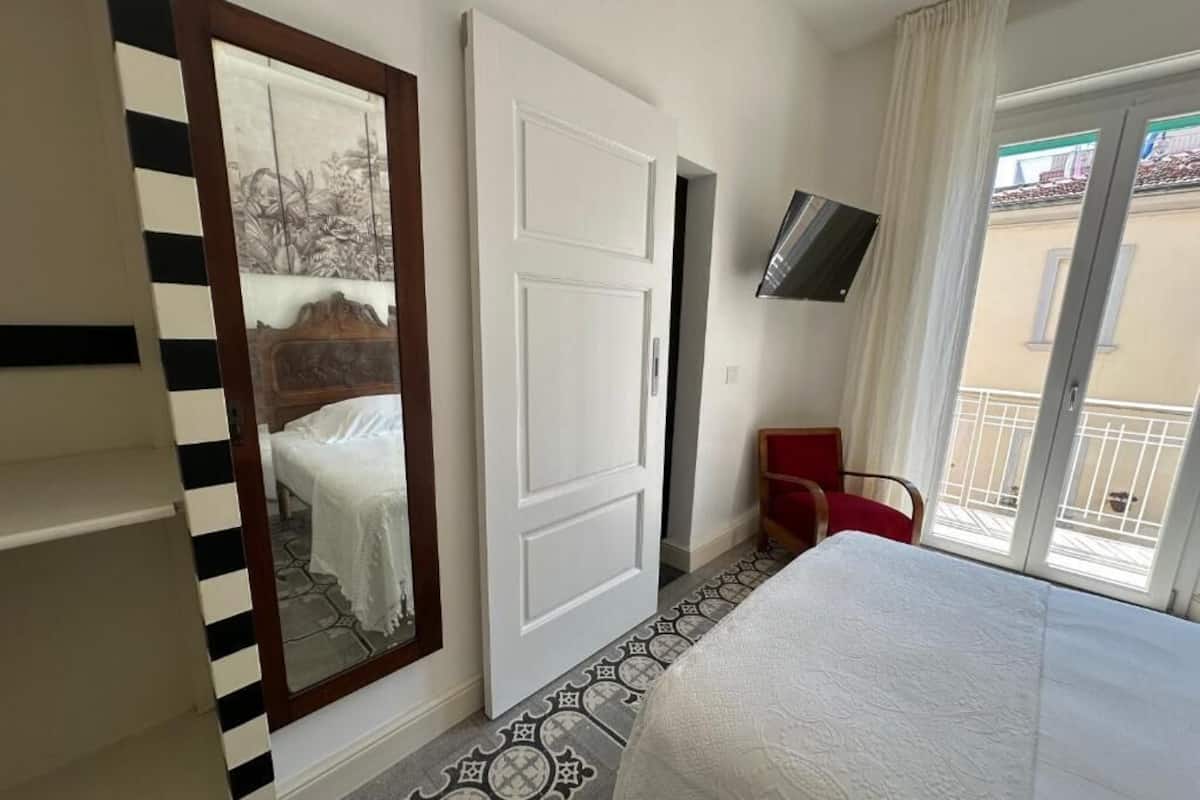 Habitación de hotel con cama, un espejo grande, armario, televisor montado en la pared y balcón con una silla roja.