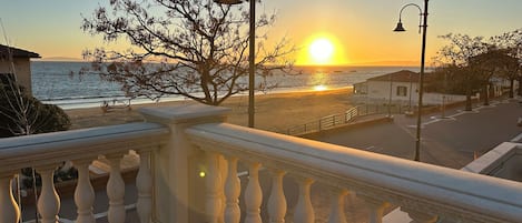 A balcony with a view of the sunset over the sea, street lamps, and a tree.