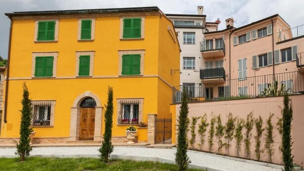 A yellow building with green shutters, a central entrance, and a small garden area with two trees. - IL GIARDINO SEGRETO (Castelfidardo)