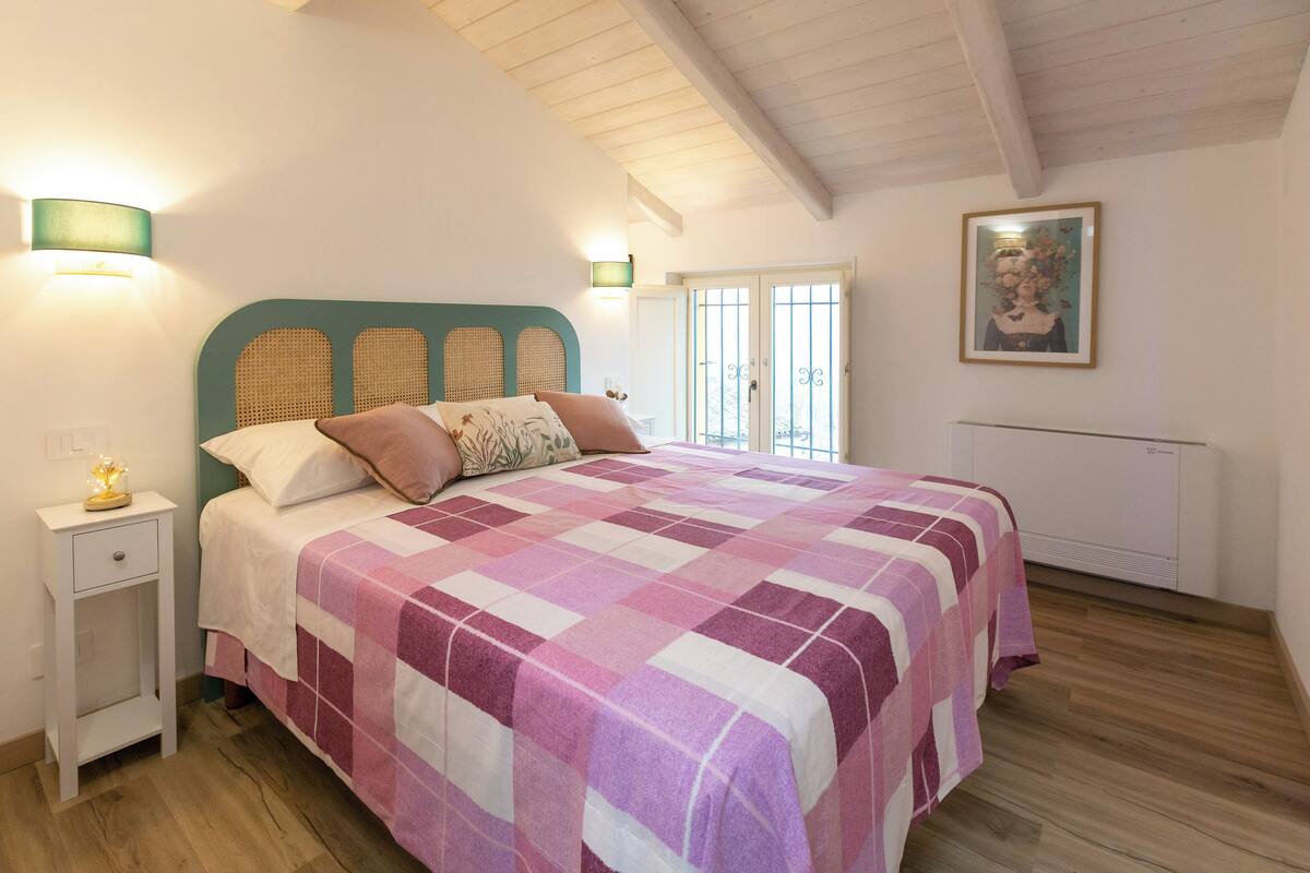 A bedroom with a checkered bedspread, a bedside table with a lamp, a window with a view, and a framed picture on the wall.