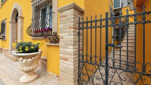 A yellow building with a wrought iron gate, a potted plant with flowers, and a brick wall. - IL GIARDINO SEGRETO (Castelfidardo)