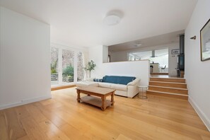 A modern living room with a wooden floor, a white sofa with a blue cushion, a wooden coffee table, and a staircase leading to another level.