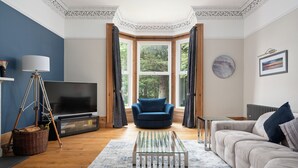 A living room with a television, a blue armchair, a sofa, and a bay window with curtains.