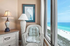 Een kamer met uitzicht op een strand, een rieten stoel, een lamp en een lijst met een afbeelding aan de muur.