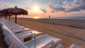 Una spiaggia con sedie a sdraio bianche e un ombrellone di paglia, un ponte sullo sfondo e un tramonto.