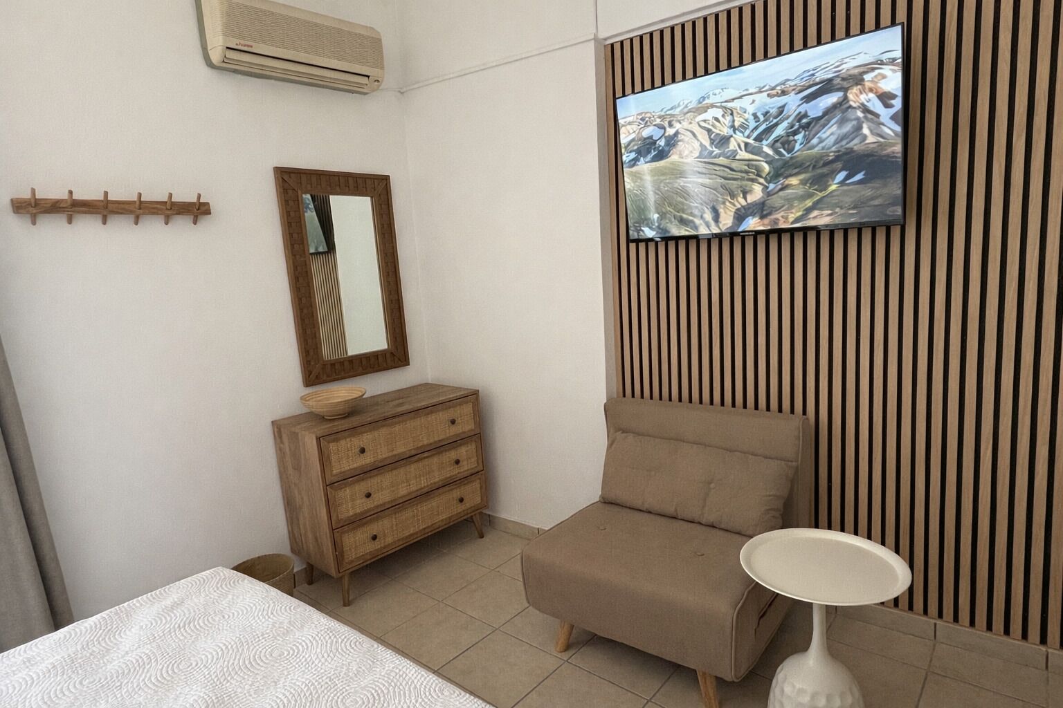 A bedroom with a bed, a wooden dresser, a chair, a TV on a wooden panel wall, and a bedside table.
