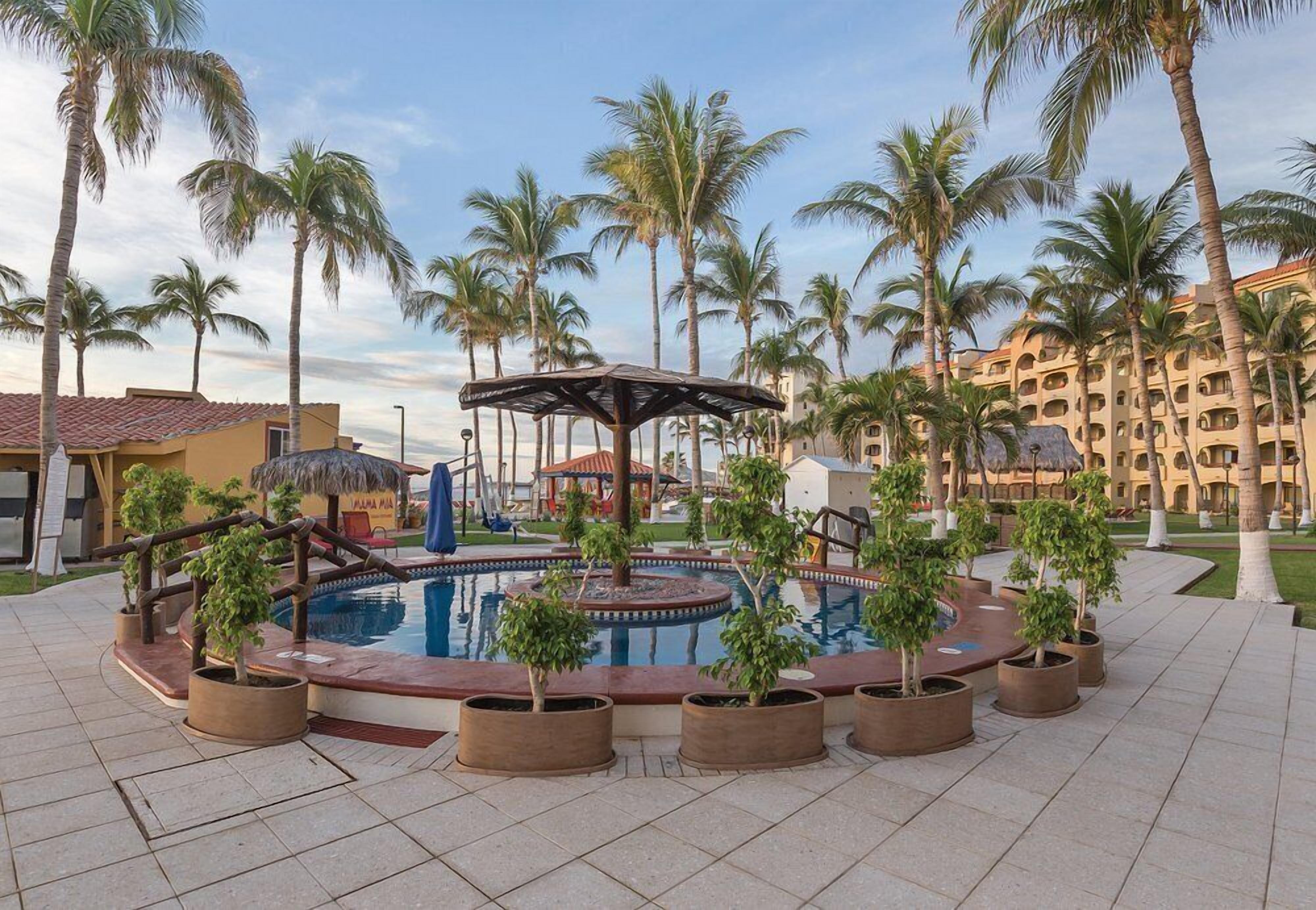 Une zone avec piscine, des palmiers, une structure à toit de chaume et, au fond, le bâtiment de l’hôtel.