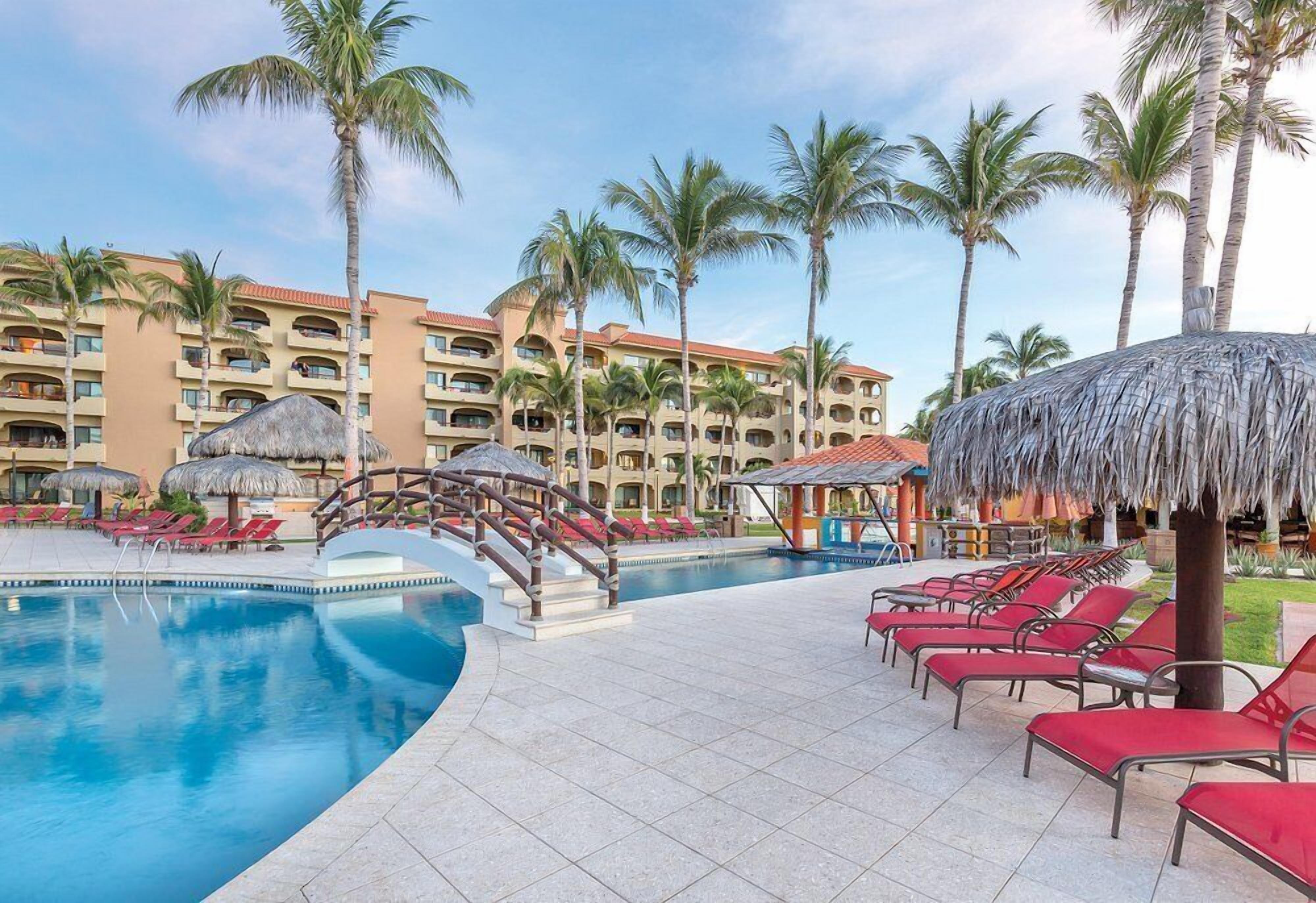 Een zwembadterras van een resort met ligbedden, palmbomen en een gebouw met balkons.