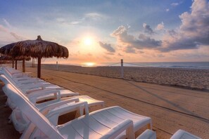 Une plage avec des chaises longues blanches et un parasol en chaume, un pont en arrière-plan et un coucher de soleil.