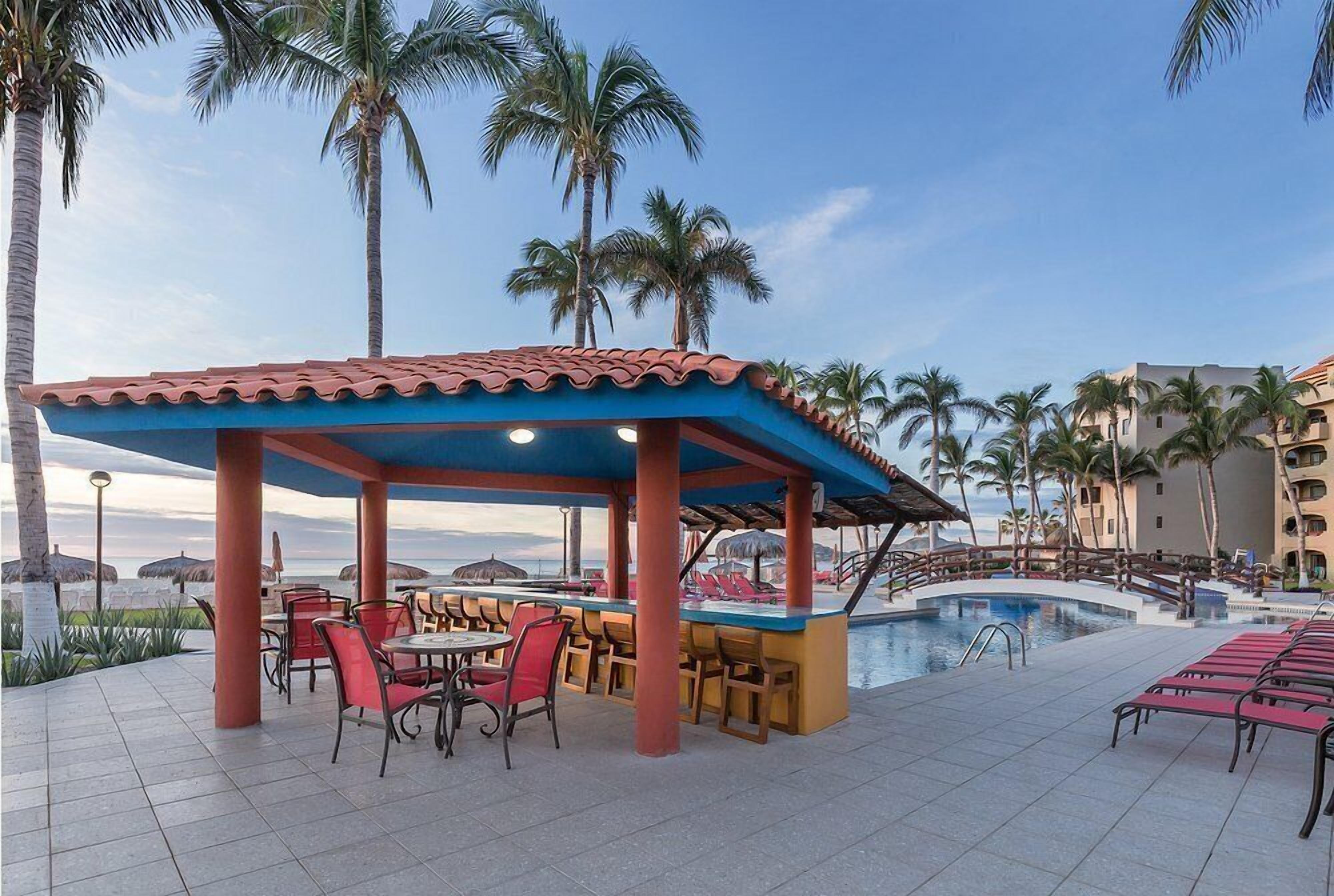 Un espace aménagé au bord de la piscine, avec un bar couvert, des sièges en plein air et des palmiers.