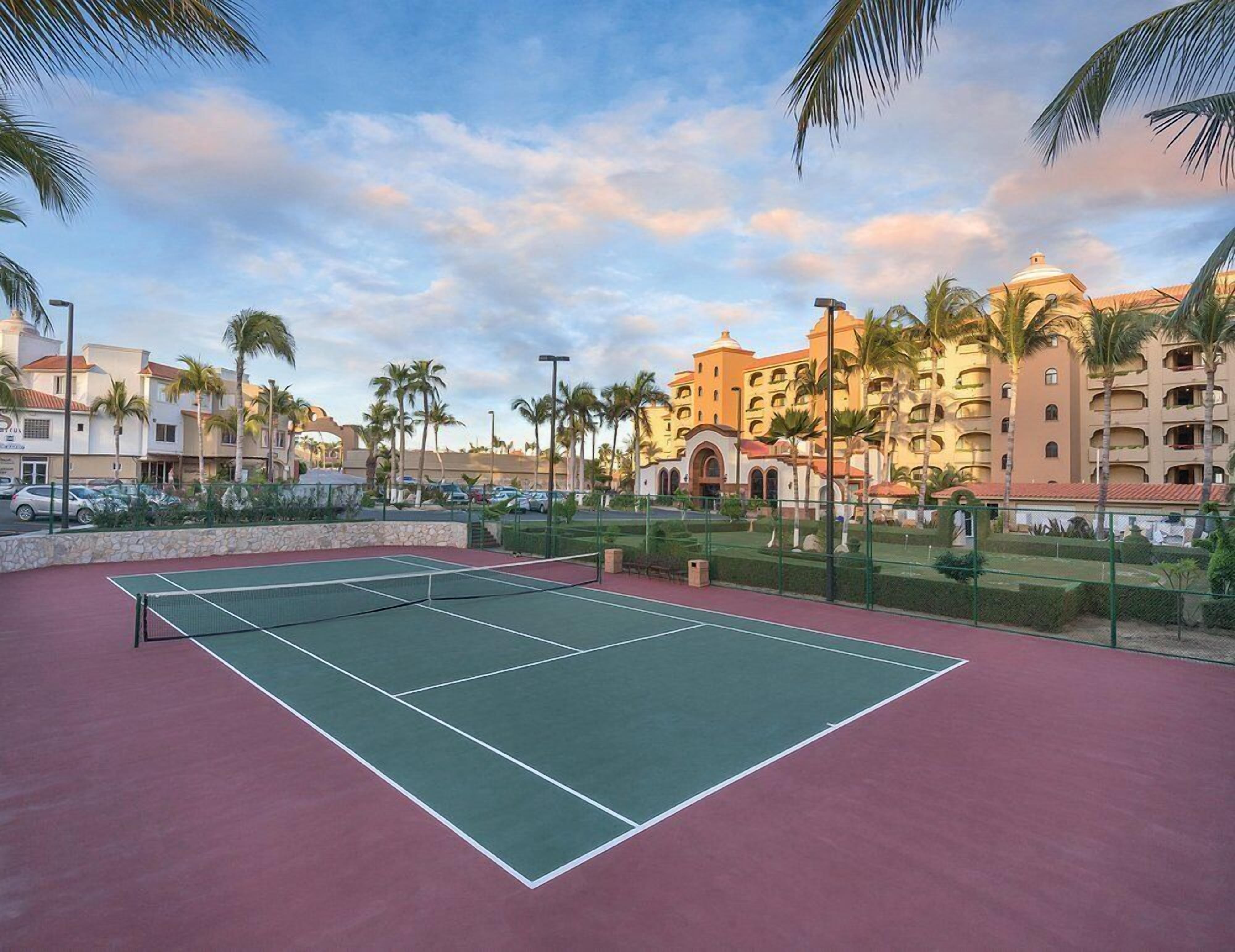 A tennis court with a net, surrounded by palm trees and a hotel with multiple balconies.
