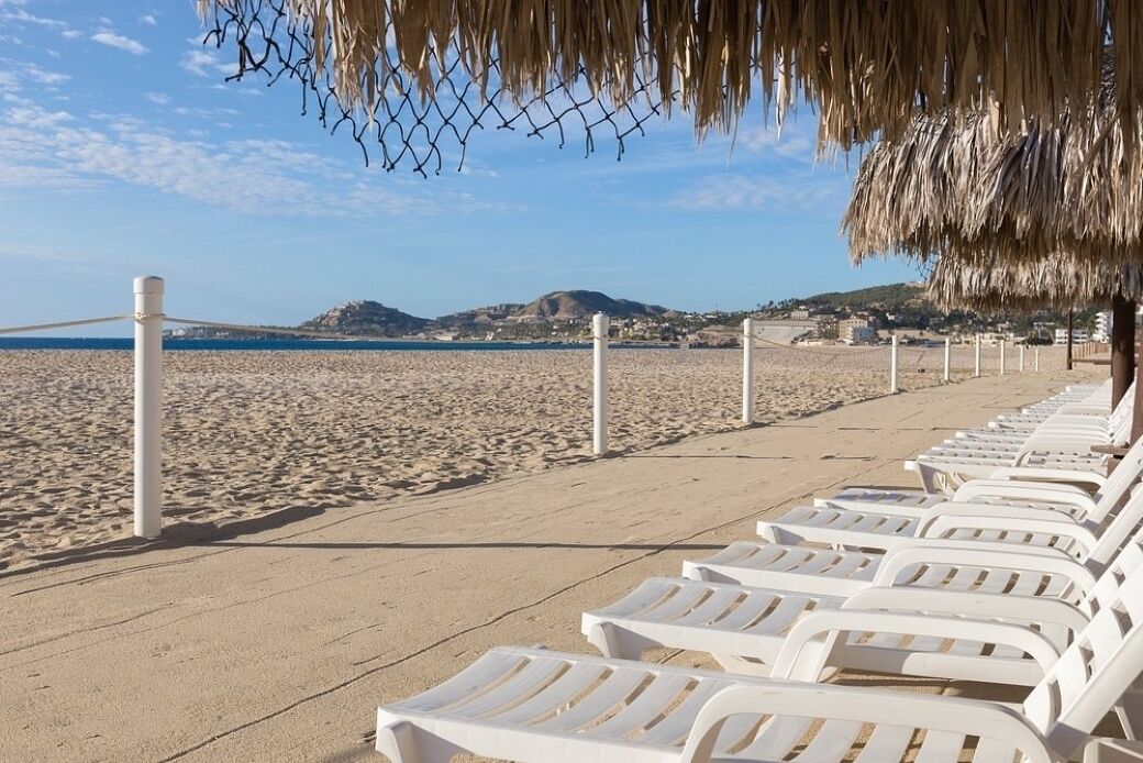 A beach with white lounge chairs lined up, a thatched roof structure, and a clear view of the sea and distant hills.