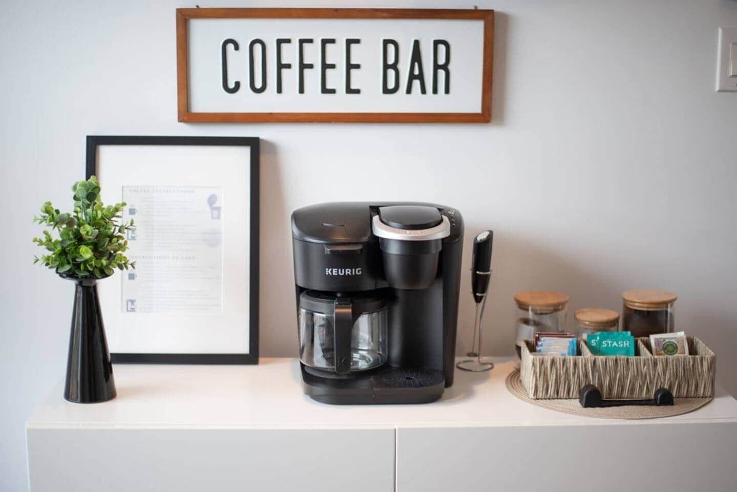 A coffee bar setup with a Keurig coffee maker, a framed menu, a small plant, and a basket with jars and a knife.