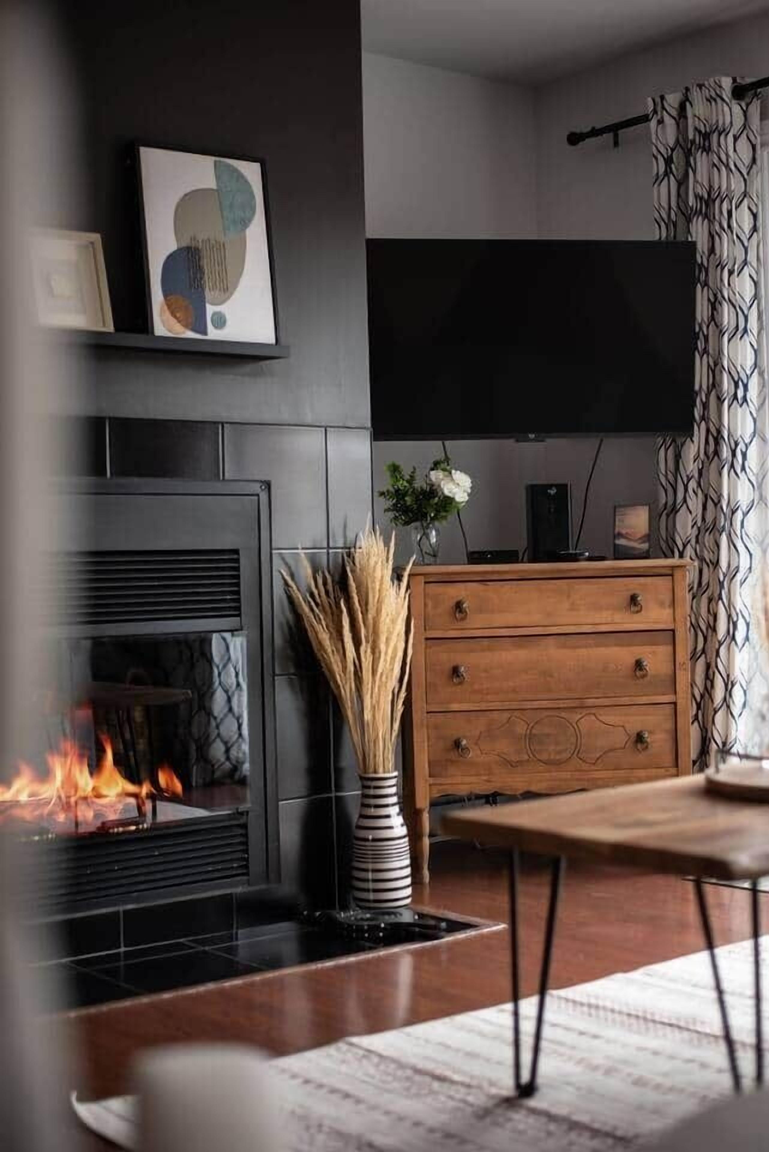 A modern living room with a fireplace, a flat-screen TV, a wooden dresser, and a vase with dried plants.