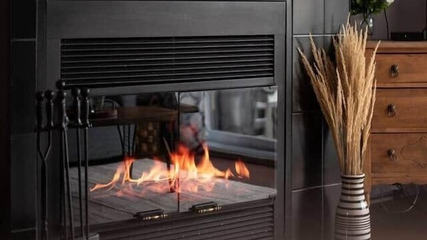 A modern fireplace with a glass front, a dark wall with a mounted shelf above it, and a wooden dresser to the right. - La Place du Village (Magog)