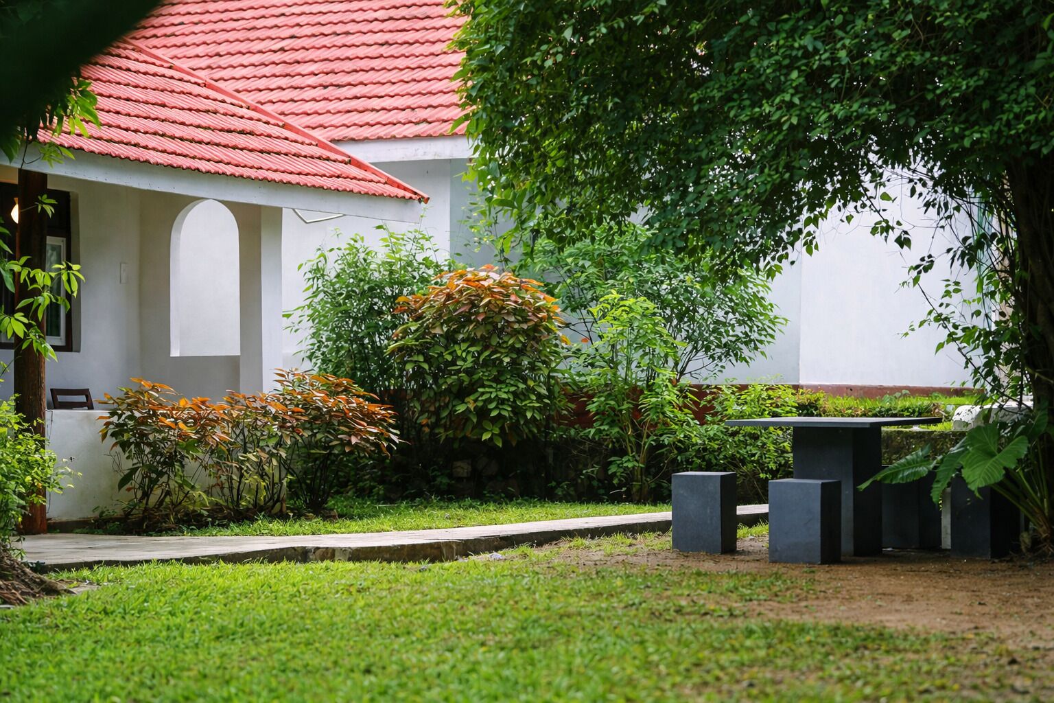 A white building with a red roof, surrounded by greenery and a small garden area.