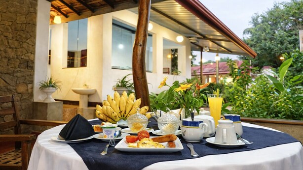 A table set for breakfast with a variety of food items, including bananas, eggs, and a glass of orange juice, in an outdoor dining area with a view of the surrounding landscape. - Habarana Inn - Offiacl (Habarana)