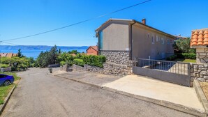 A residential street with a house featuring a stone facade, a gated driveway, and a view of the sea. - Cozy apartment for 2 people in Novi Vinodolski with balcony and barbecue (Novi Vinodolski)