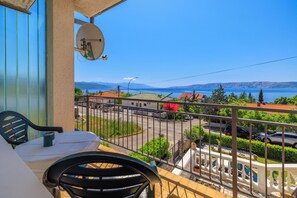 A balcony with a table and chairs, a satellite dish, and a view of a lake and buildings. - Cozy apartment for 2 people in Novi Vinodolski with balcony and barbecue (Novi Vinodolski)