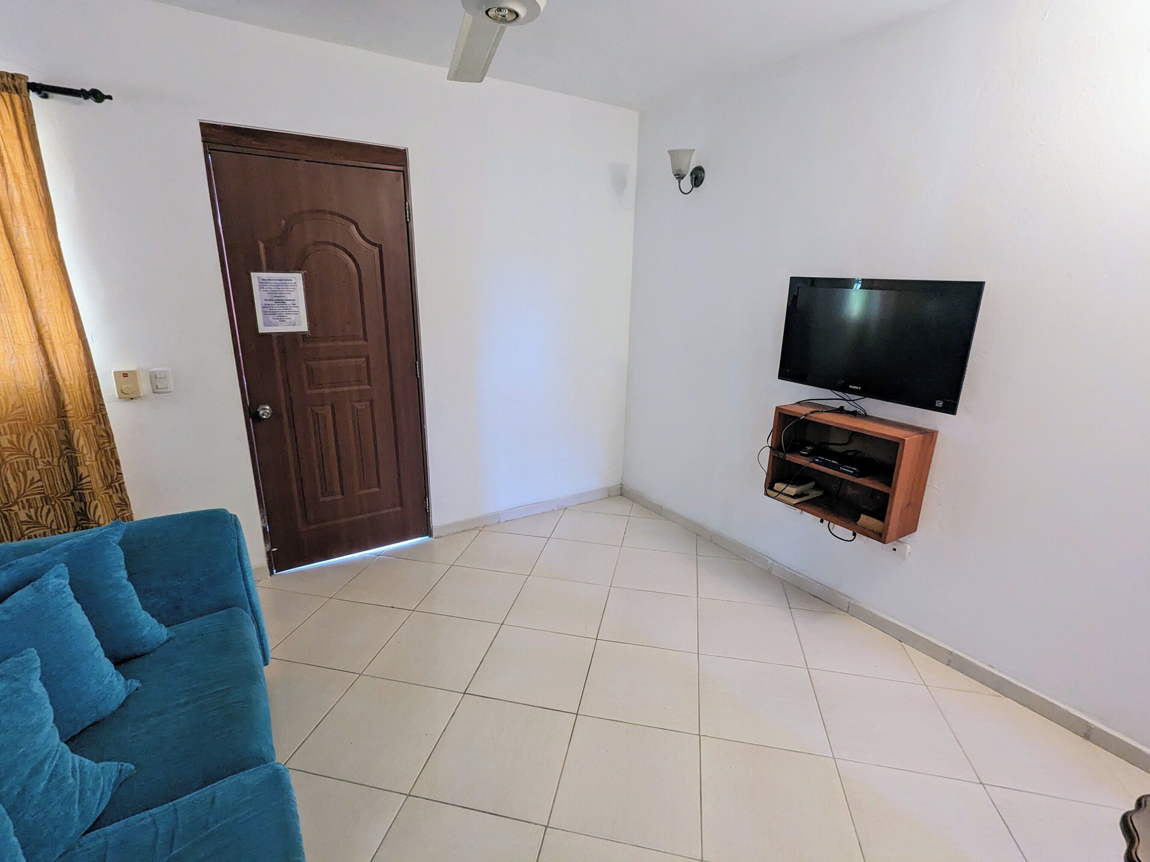 A room with a wooden door, a flat-screen TV mounted on the wall, a blue sofa, and a tiled floor.