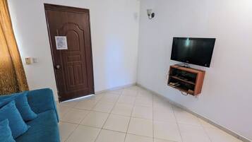A room with a wooden door, a flat-screen TV mounted on the wall, a blue sofa, and a tiled floor.