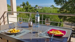 Una mesa de comedor con platos de comida, copas de vino y botellas en un balcón con vistas a zonas verdes y edificios.