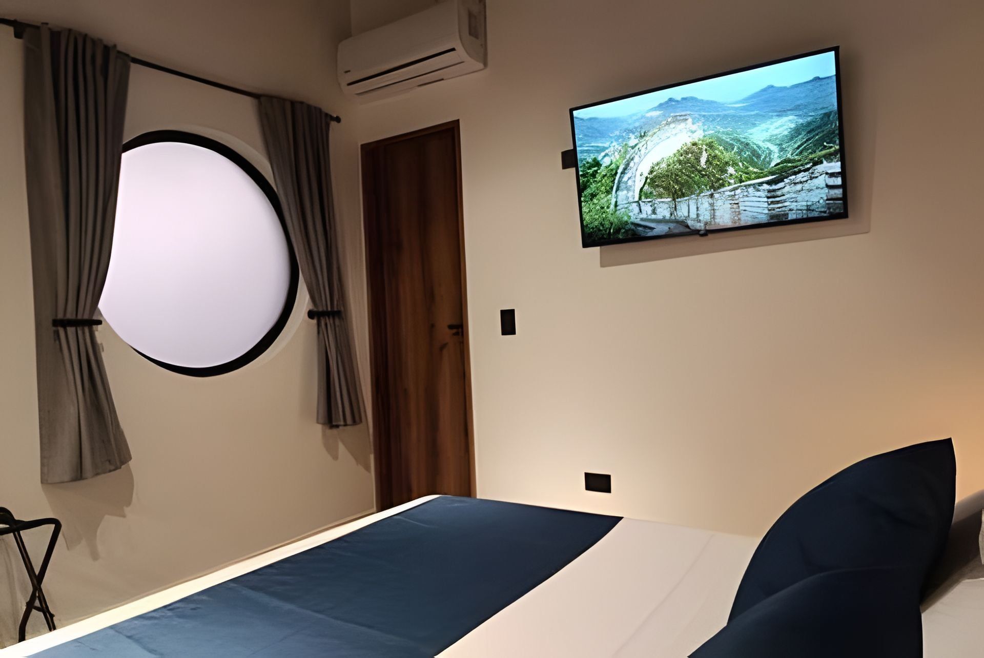 A bedroom with a bed, a mounted TV displaying a scenic landscape, a circular light fixture, and a door.