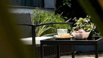 A table with a bowl of fruit and a cup, set on a balcony with a view of greenery and a building in the background.