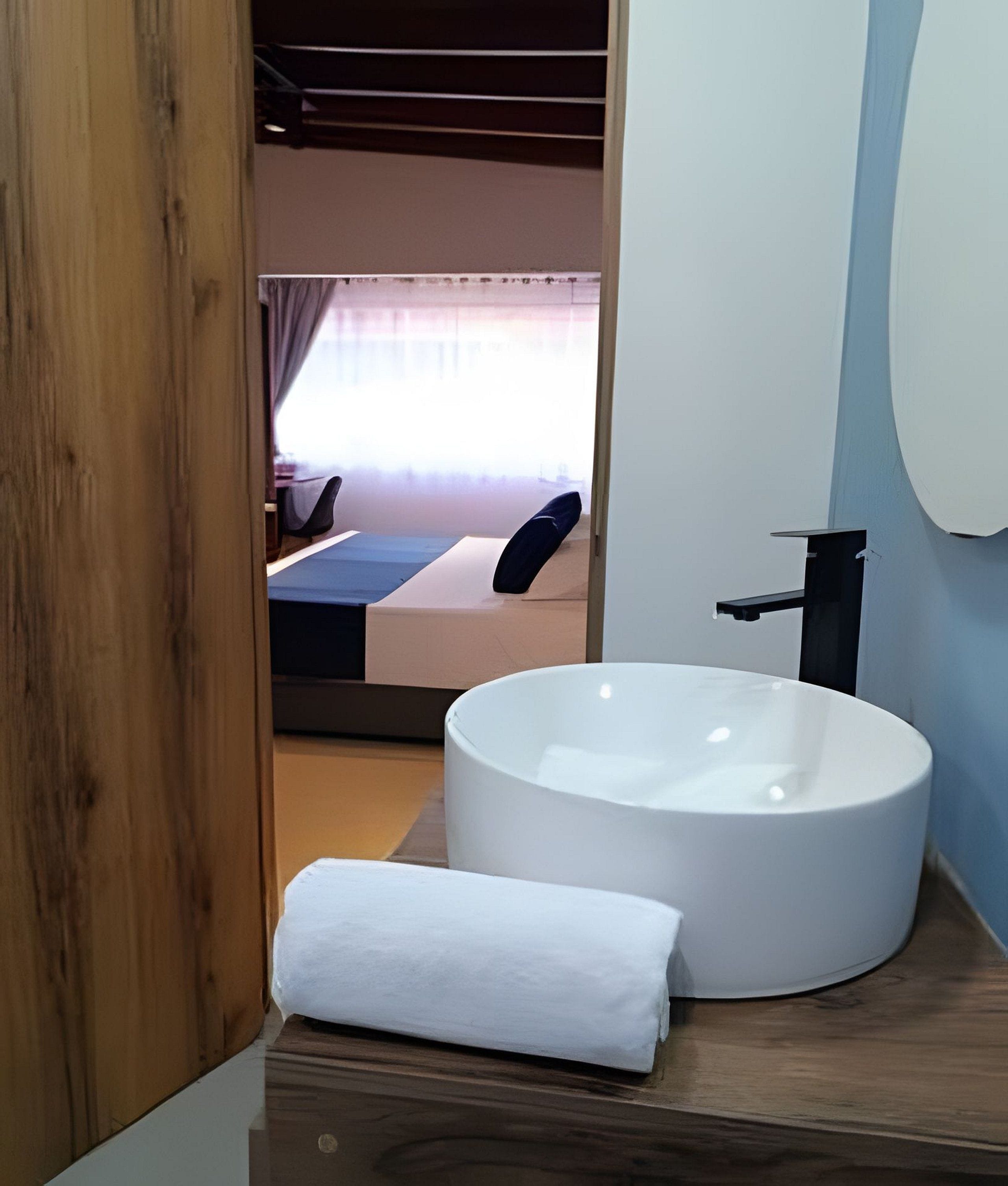 A modern bathroom with a large white bathtub, a wooden floor, and a visible bedroom with a bed and curtains in the background.