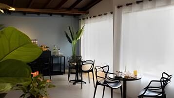 Un coin repas avec une table dressée pour deux personnes, entourée de chaises, d’une grande plante en pot et d’une fenêtre avec des rideaux.