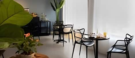 Un área de comedor con una mesa dispuesta para dos, rodeada de sillas, una planta grande en maceta y una ventana con cortinas.