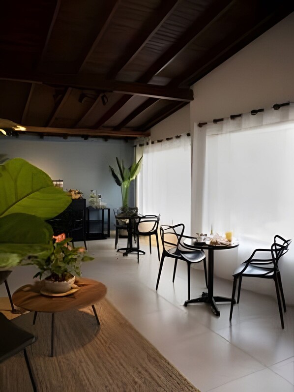 A dining area with a table set for two, surrounded by chairs, a large potted plant, and a window with curtains.