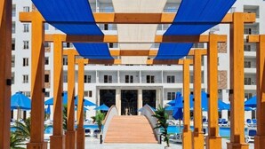 Een hotelentree met een houten boog, een zwembadgedeelte met parasols, en een gebouw met meerdere verdiepingen op de achtergrond.