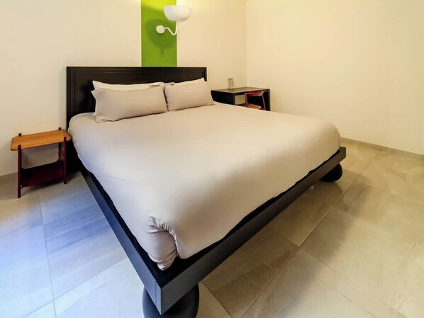Una cama con estructura negra, una colcha blanca y dos almohadas. Hay una mesita de noche de madera con una lámpara encima. La habitación tiene una pared de acento verde y un techo blanco.