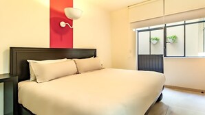 A neatly made bed in a modern hotel room with a red accent wall, a bedside table, and a window with potted plants. - Apt 2BR balcony, terrace, Heart of Guadalajara (Guadalajara)