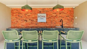 Eine Bar mit einer Ziegelwand, einem Schild mit der Aufschrift 'ROMA 1888' und vier grünen Stühlen.