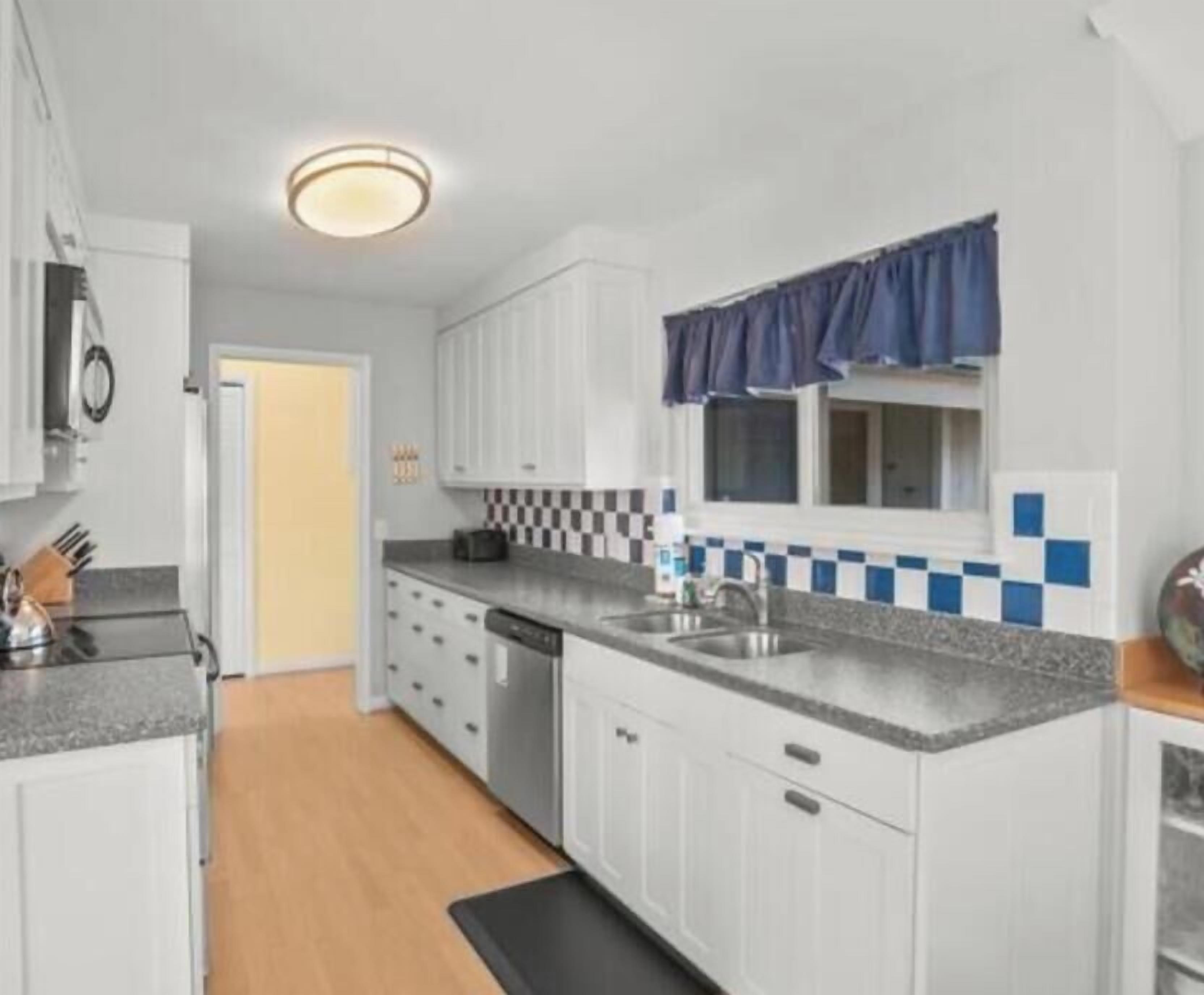 A compact kitchen with white cabinets, a granite countertop, a dishwasher, and a window with a blue valance.